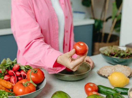 Person in pink overshirt holding a fresh tomato with various organic vegetables on a countertop.