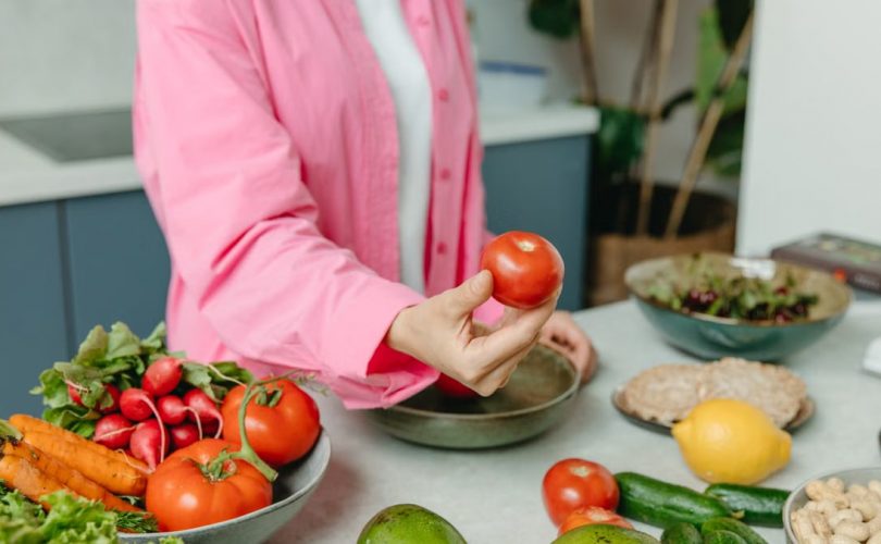 Person in pink overshirt holding a fresh tomato with various organic vegetables on a countertop.