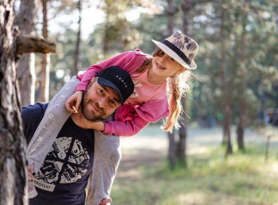 A joyful father giving his daughter a piggyback ride in a sunny forest setting.