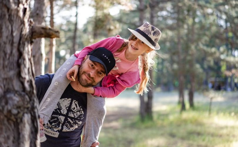 A joyful father giving his daughter a piggyback ride in a sunny forest setting.